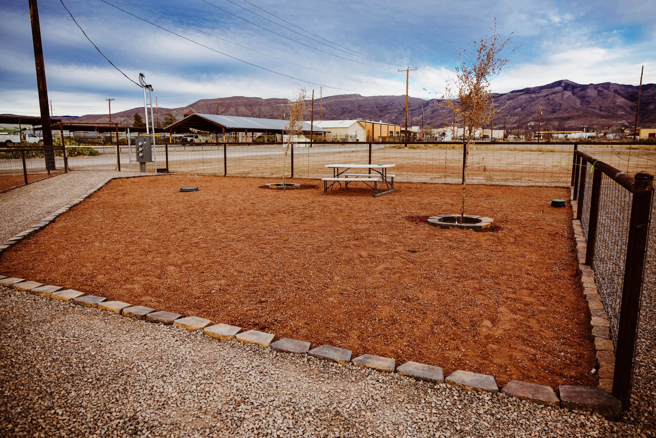 A fenced-in dirt yard with a picnic table and two small trees. Mountains are visible in the background.