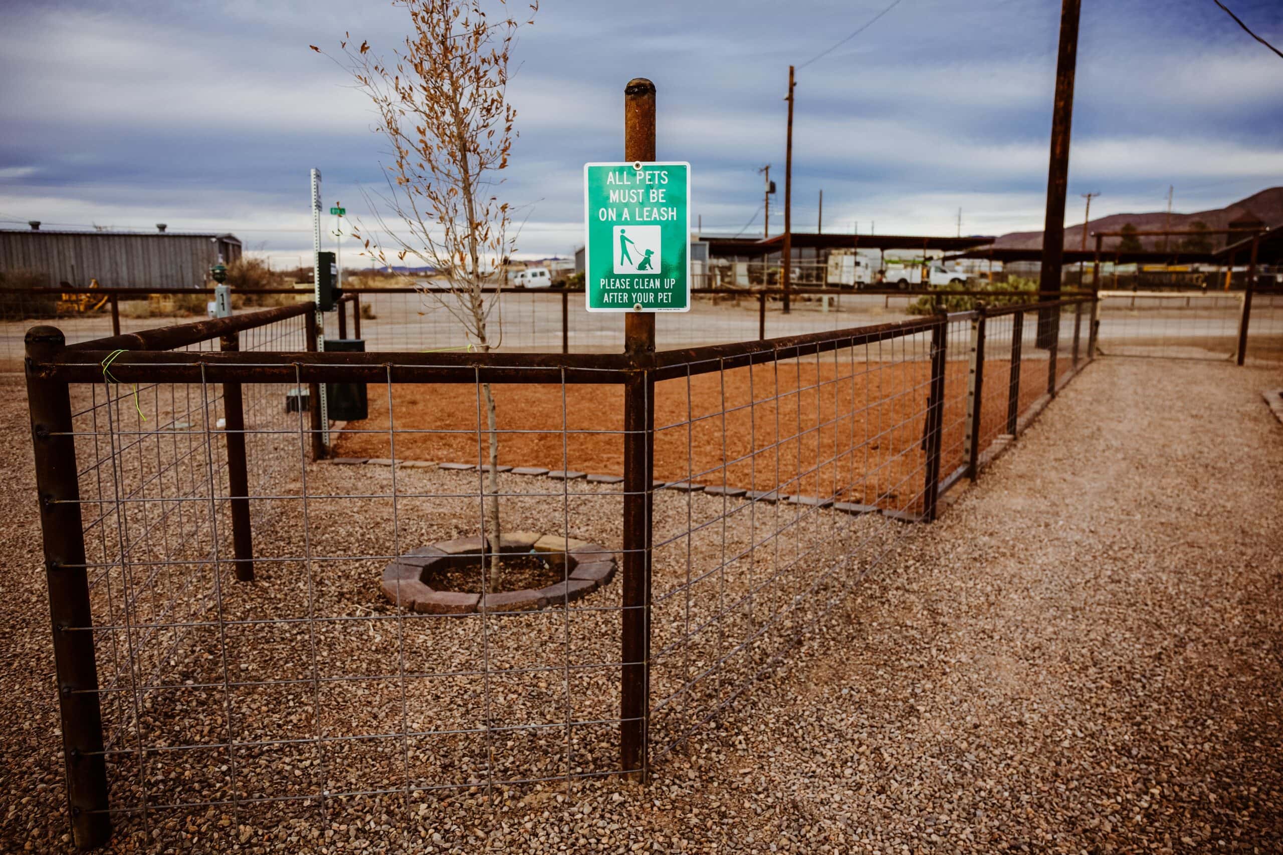 Fenced dog area with a sign that reads "All pets must be on a leash." Gravel path and a tree visible inside the fenced space.